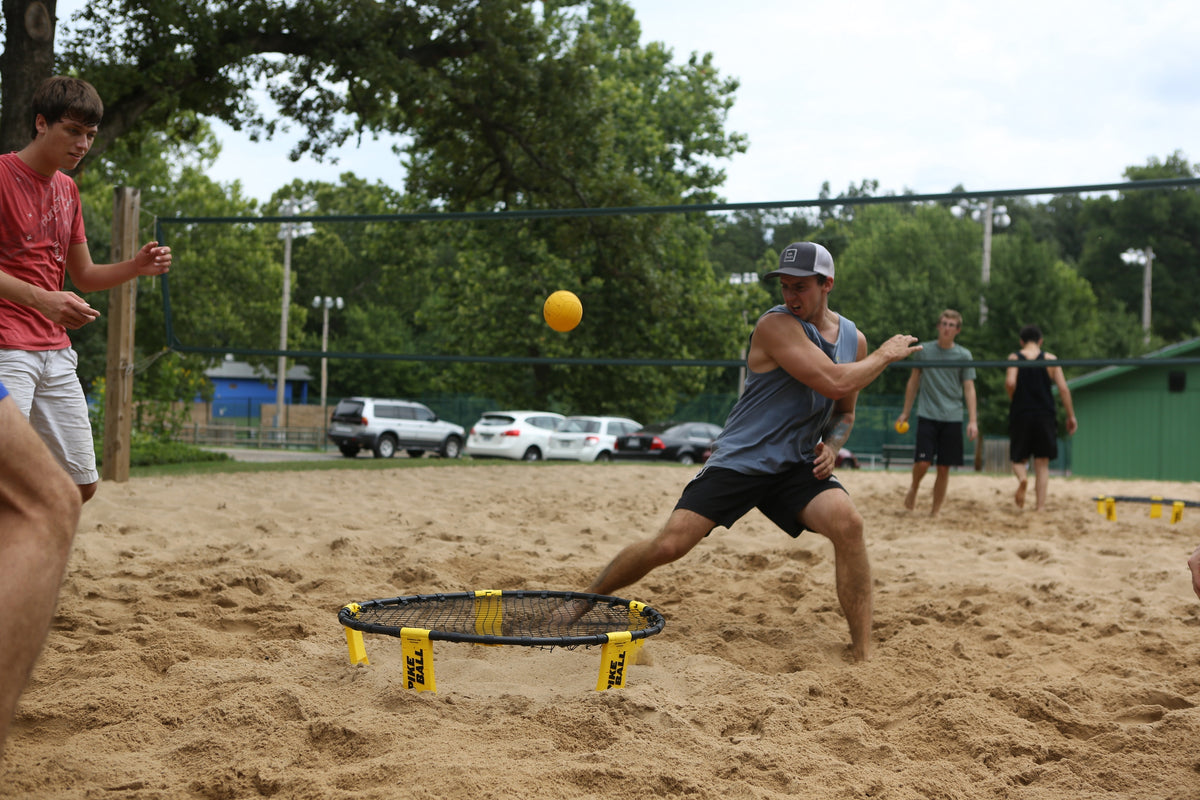 Four young men play Spikeball on a sandy court, with one in a blue tank top lunging to hit the yellow ball on a Kankakee Spikeball Standard Set featuring an adjustable net. Trees, cars, and a green building are visible in the background.