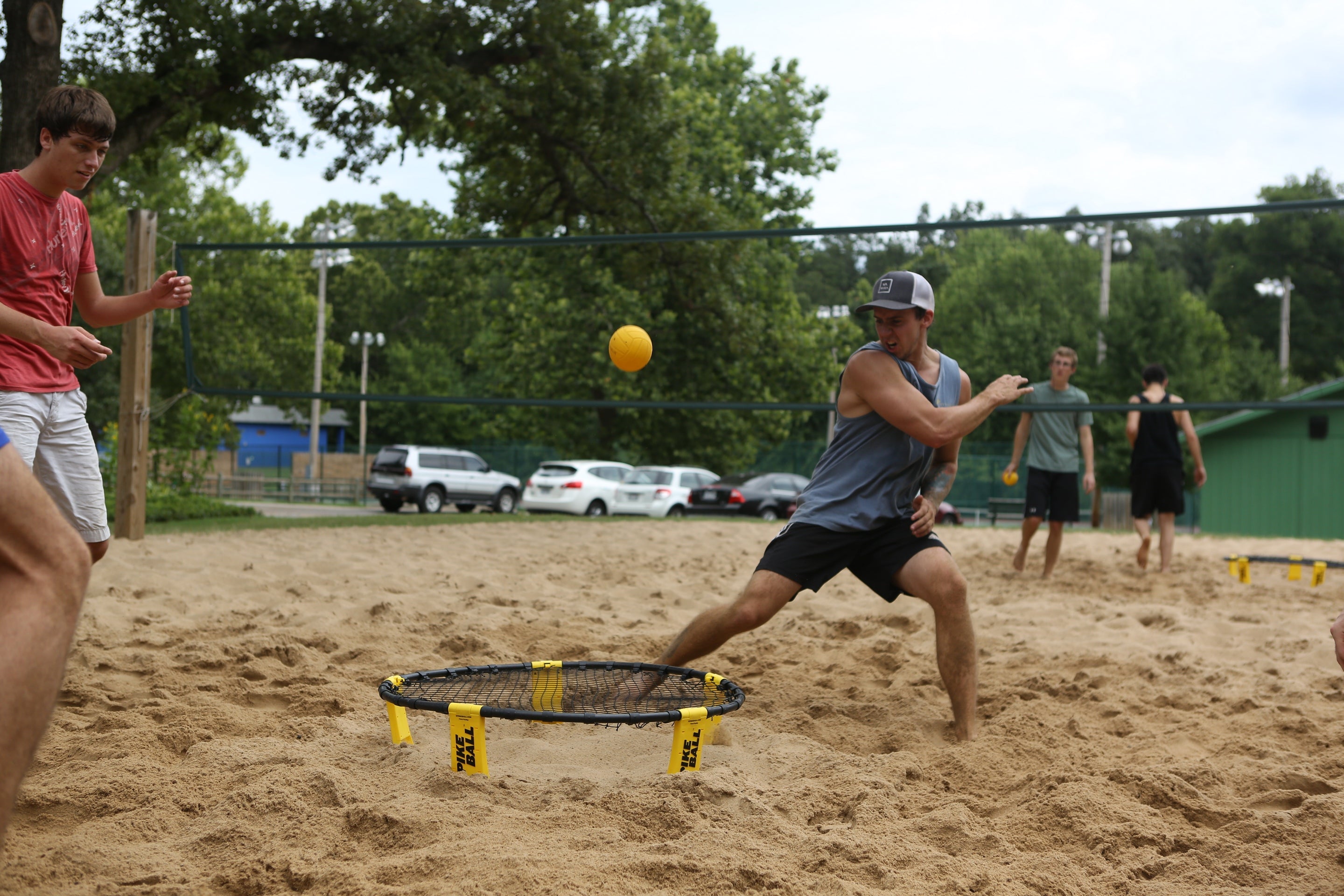 Four young men play Spikeball on a sandy court, with one in a blue tank top lunging to hit the yellow ball on a Kankakee Spikeball Standard Set featuring an adjustable net. Trees, cars, and a green building are visible in the background.