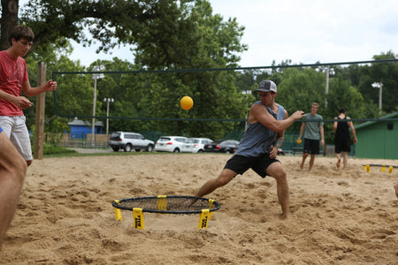 Four young men play Spikeball on a sandy court, with one in a blue tank top lunging to hit the yellow ball on a Kankakee Spikeball Standard Set featuring an adjustable net. Trees, cars, and a green building are visible in the background.