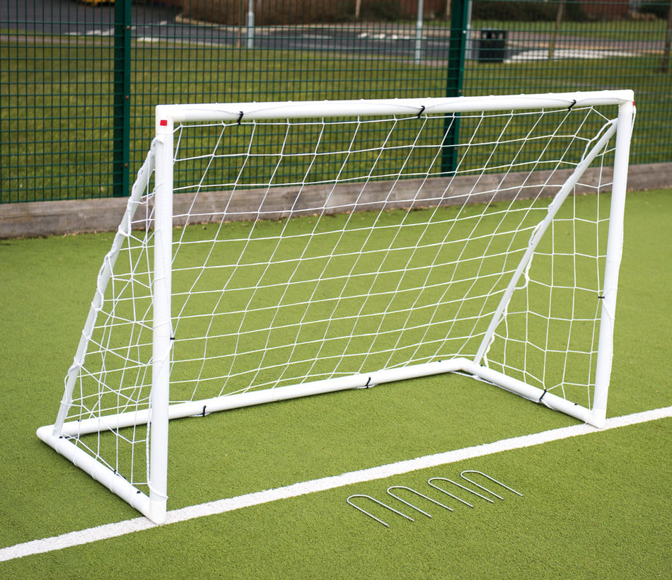 The Precision Training Precision Junior Garden Goal (8' x 6') with a white plastic frame and net is set up on green artificial turf. Four ground anchors are in front, with a fence and outdoor area visible in the background.