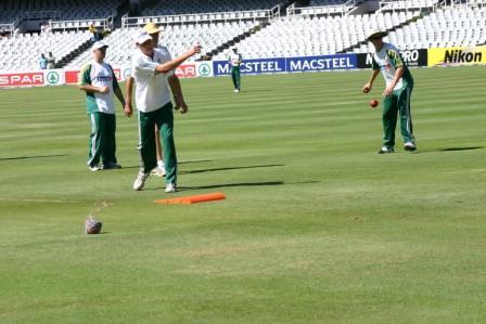 Four people in sports gear practice cricket on a grassy field. One throws a ball toward the stumps while others watch and use the Katchet Training Aid by Katchet for catching drills. The stadium seats behind them are empty.