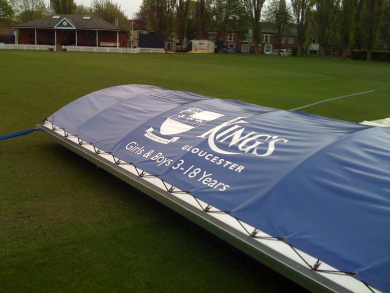 A large blue and white Ultimate Mobile Cricket Pitch Cover by Martin Berrill Sports is set on a sports field. The cover displays “KING’S GLOUCESTER Girls & Boys 3-18 Years,” with trees and buildings visible in the background.