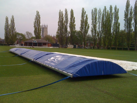 The Martin Berrill Sports Ultimate Mobile Cricket Pitch Covers, in blue and white, are set on a grassy field with tall trees and a church tower in the cloudy background.