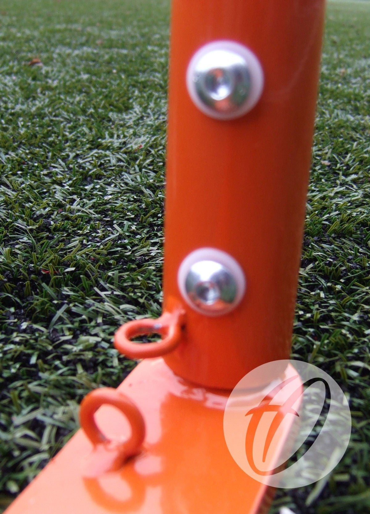 Close-up of an orange metal pole with bolts and two circular hooks, part of Harrod Sport Freestanding Competition Lacrosse Goals on artificial green turf. A semi-transparent stylized W logo appears in the image's bottom right corner.