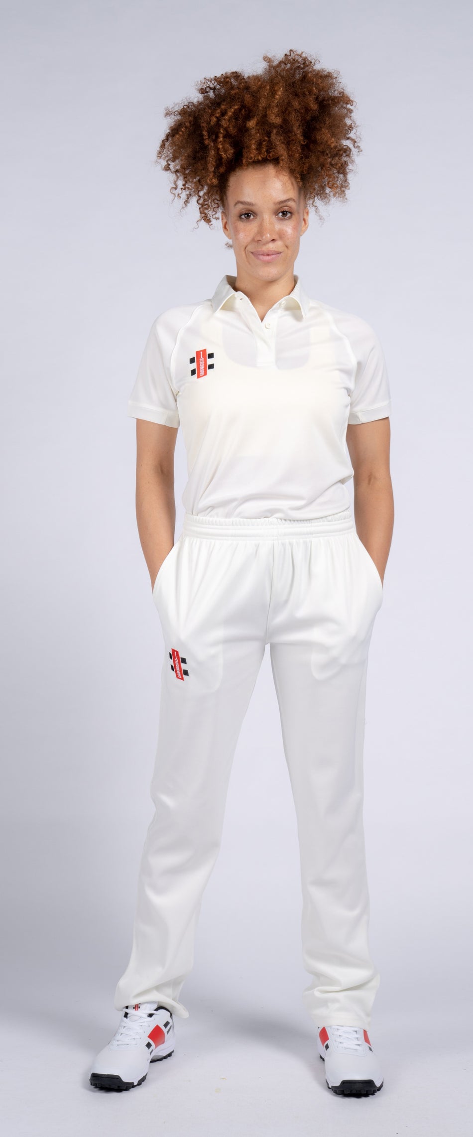 A person with curly hair wears Gray Nicolls Matrix V2 Ladies Trousers and white sneakers, standing forward with hands in pockets, dressed in a white cricket uniform against a plain light background.