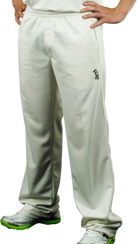 A person stands hands on hips, showing only their lower body dressed in off-white Kookaburra Pro Players Trousers Senior, a white shirt, and white athletic shoes with green soles.