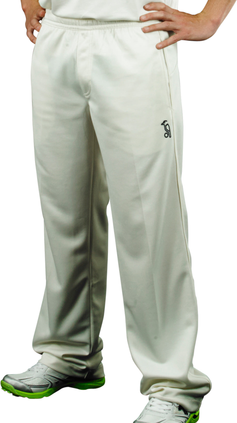 A person stands hands on hips, showing only their lower body dressed in off-white Kookaburra Pro Players Trousers Senior, a white shirt, and white athletic shoes with green soles.