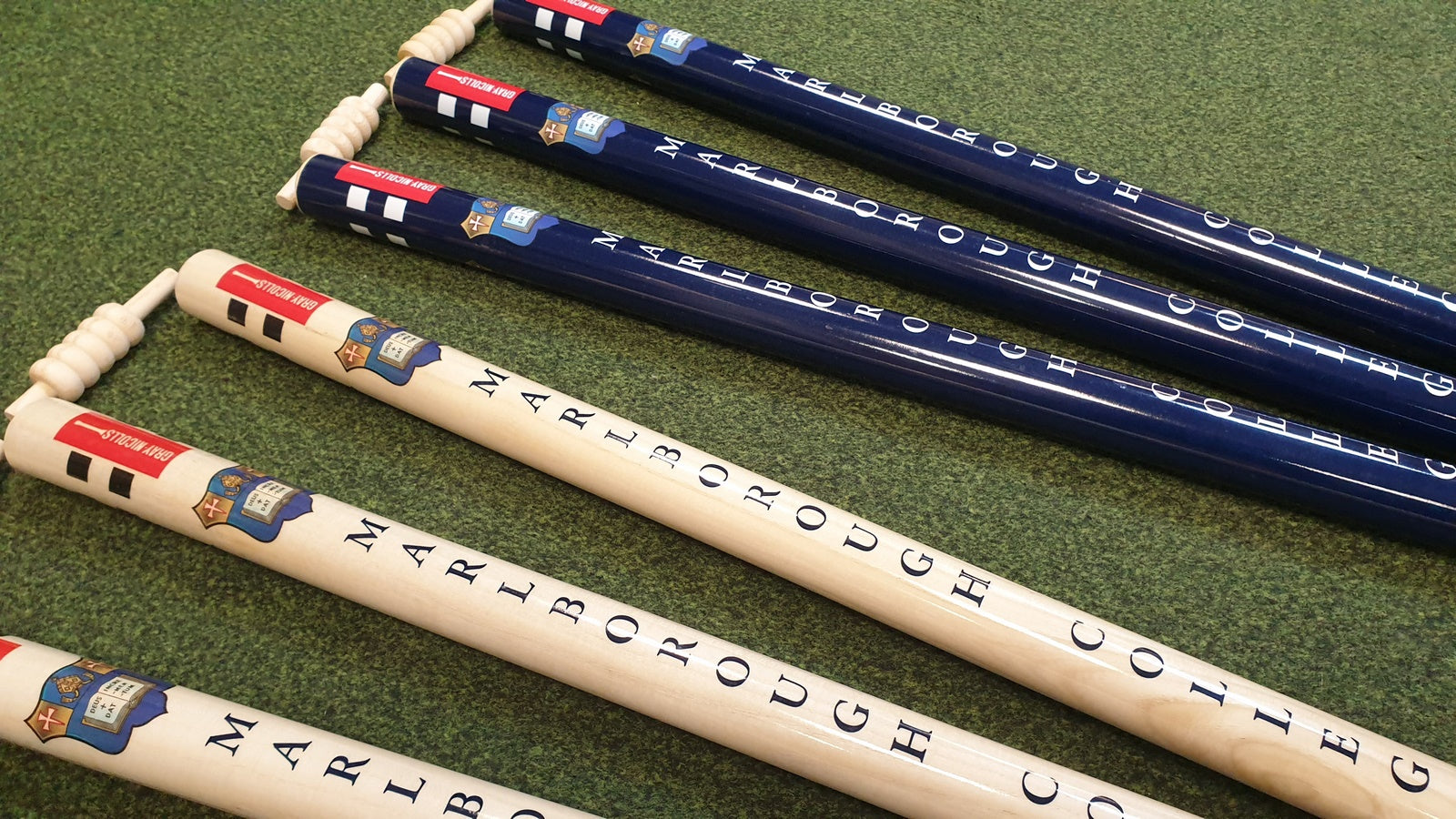 Five croquet mallets with Marlborough College on the handles—three dark blue, two white—rest on green grass. Nearby, Gray Nicolls Custom Printed Club Cricket Stumps add a personalised touch to the scene.