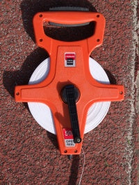 A Martin Berrill Sports Measuring Tape 50m with orange fibreglass reel, black handle and crank, lies on a textured reddish outdoor surface, the tape partially extended.
