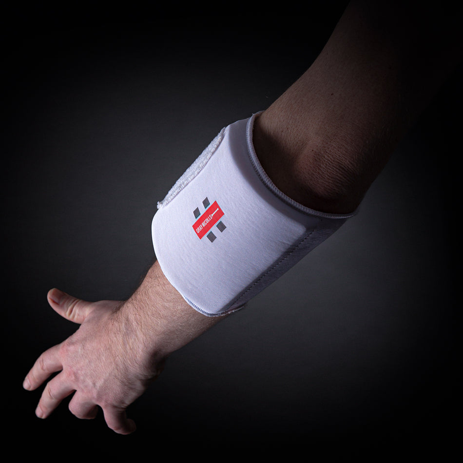 An arm wearing the Gray Nicolls Mini Pro Armguard, featuring a white pad with a red and black Gray Nicolls logo for padded protection, is shown against a dark background.