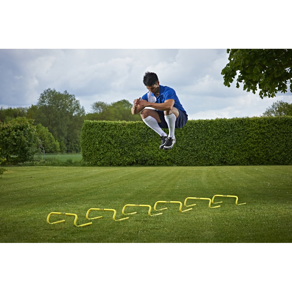Wearing athletic gear, a person jumps over Mitre Training Hurdle Set 6" on a grassy lawn, showcasing speed and power, with trees and bushes in the background.