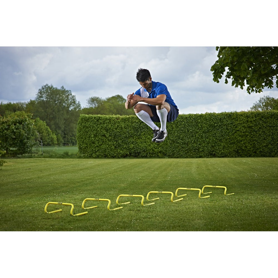 Wearing athletic gear, a person jumps over Mitre Training Hurdle Set 6" on a grassy lawn, showcasing speed and power, with trees and bushes in the background.