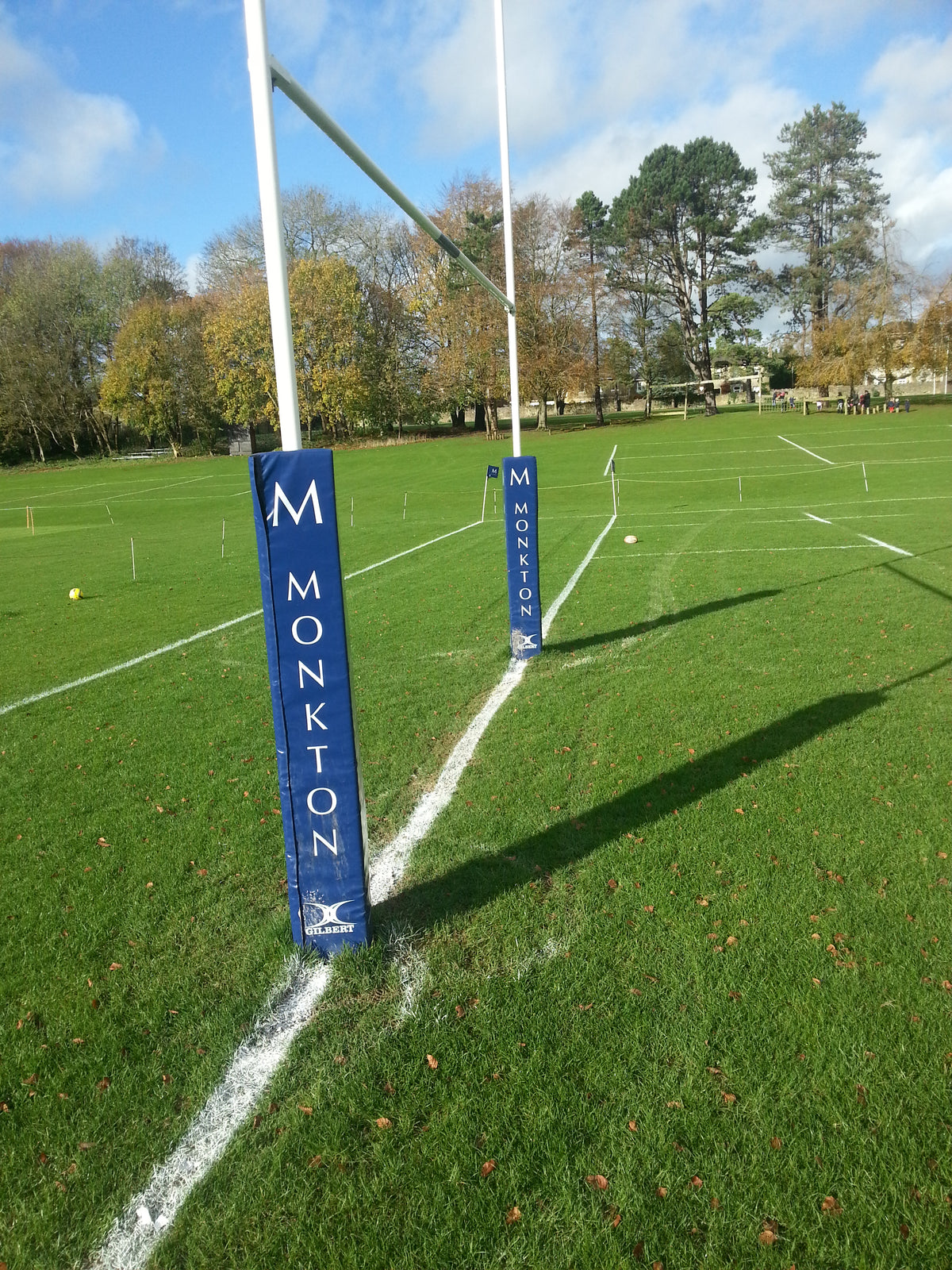Gilbert Custom Printed Senior Rugby Post Protectors, featuring the Monkton label in blue, are fitted on rugby goalposts on a green field with white pitch lines and trees in the background beneath a partly cloudy sky.