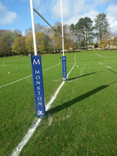 Gilbert Custom Printed Senior Rugby Post Protectors, featuring the Monkton label in blue, are fitted on rugby goalposts on a green field with white pitch lines and trees in the background beneath a partly cloudy sky.