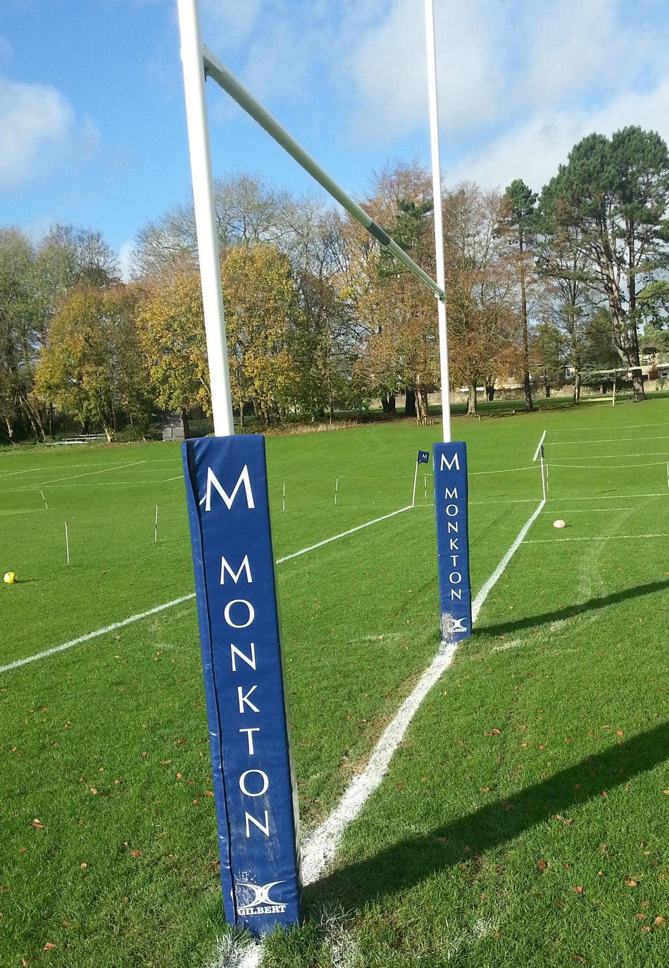 A rugby goalpost on grass features Gilbert Custom Printed Senior Rugby Post Protectors labeled Monkton. White field lines, a rugby ball, and autumn trees appear under a partly cloudy sky.