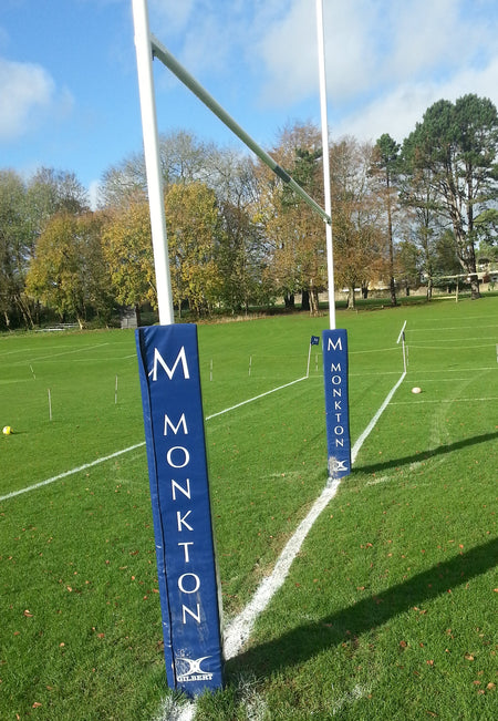 A set of Gilbert Custom Printed Junior Rugby Post Protectors on a grassy field with white lines and a rugby ball. The blue pads are labeled MONKTON, with trees and autumn leaves in the background under a partly cloudy sky.