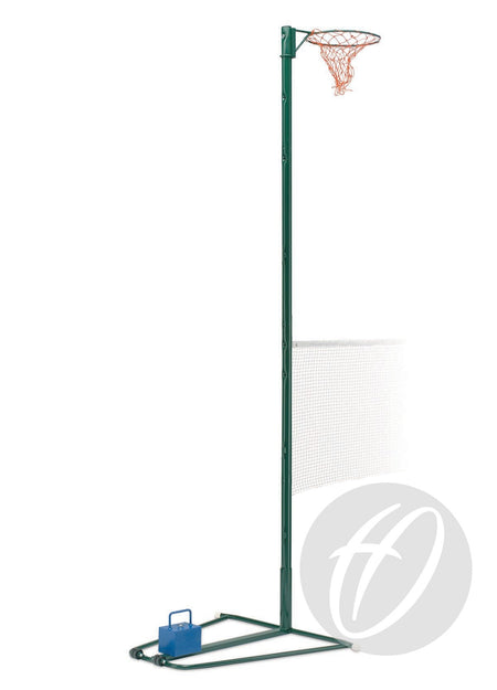 The Harrod Sport NB1AC Combination Post is a green metal netball post with an orange net and weighted base. A partial white mesh net for badminton or mini tennis and a blue weight on the base are also visible behind the post.