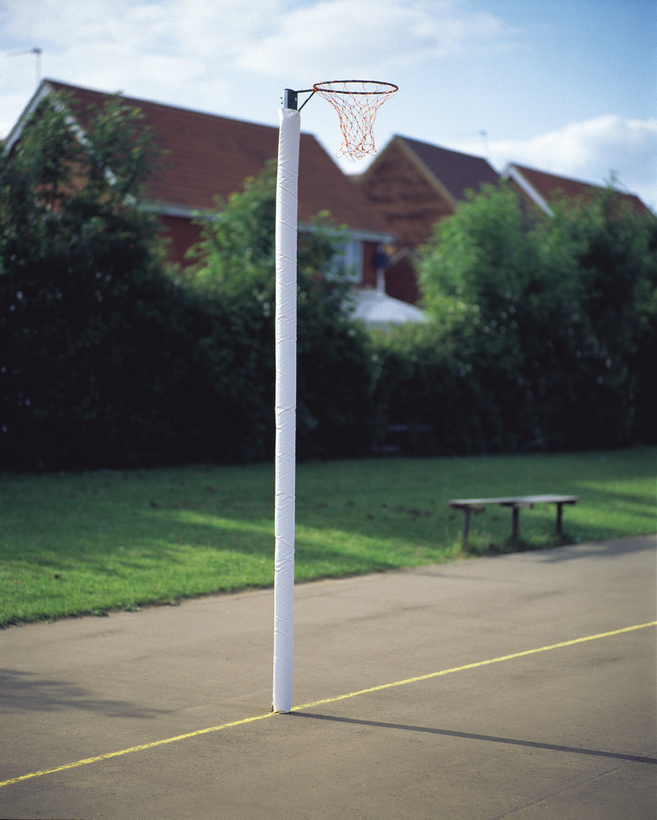 A Harrod Sport Netball Post - Socketed, 10MM Ring with a red net and adjustable ring height stands on an outdoor court, surrounded by green grass and trees, with houses featuring red roofs in the background. A bench is near the socketed posts.