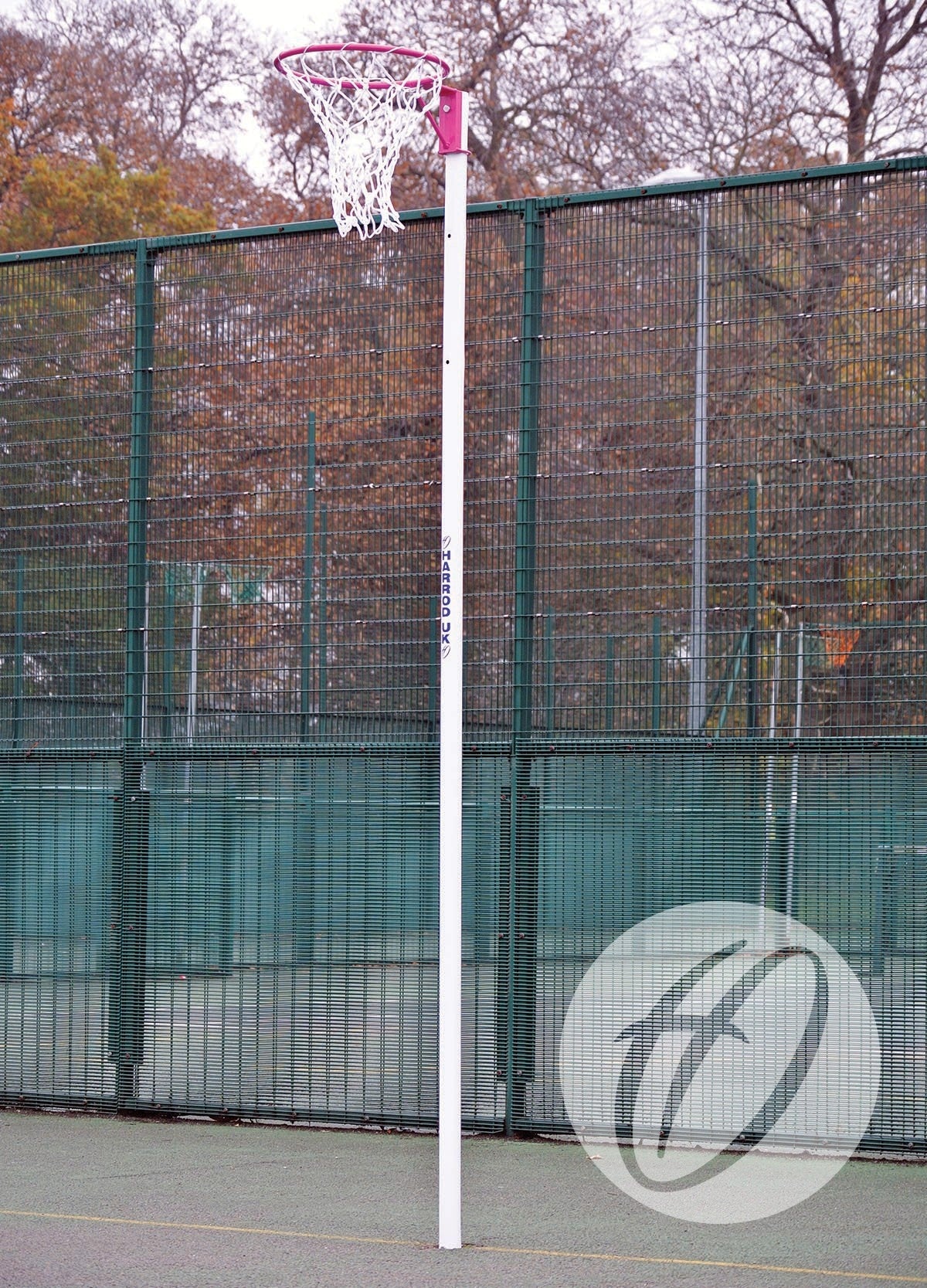 A pair of Harrod Sport Practice White Netball Nets are set up on an outdoor court with regulation-size hoops, framed by a green wire fence and autumn trees in the background.