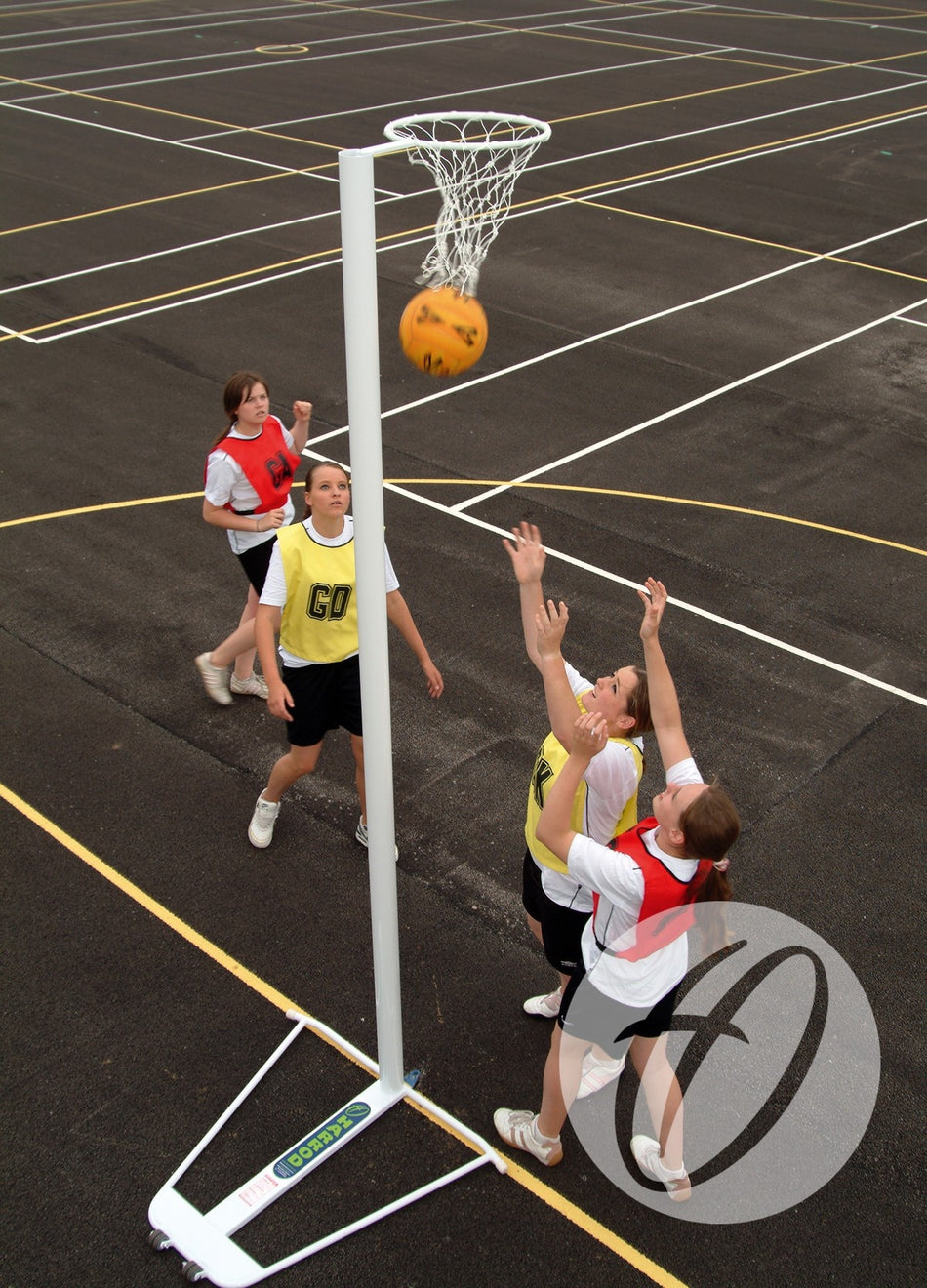 Four players on an outdoor court watch the ball pass through a Harrod Sport Practice White Netball Net, with two in yellow bibs and two in red bibs reaching up amid painted lines around the regulation hoop.