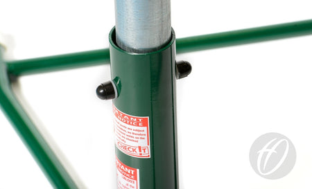 Close-up of a Harrod Sport Regulation Netball Post—Freestanding, 16MM Ring PAIR—showing a metal pole in a green frame with black locking pins, red and white warning labels, and the gray logo in the bottom right corner.