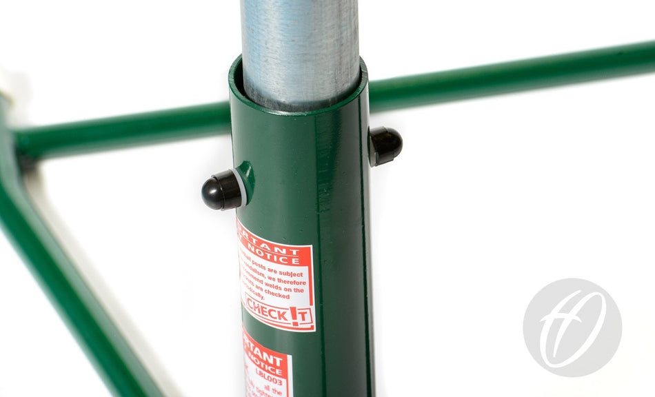 Close-up of a Harrod Sport Regulation Netball Post—Freestanding, 16MM Ring PAIR—showing a metal pole in a green frame with black locking pins, red and white warning labels, and the gray logo in the bottom right corner.