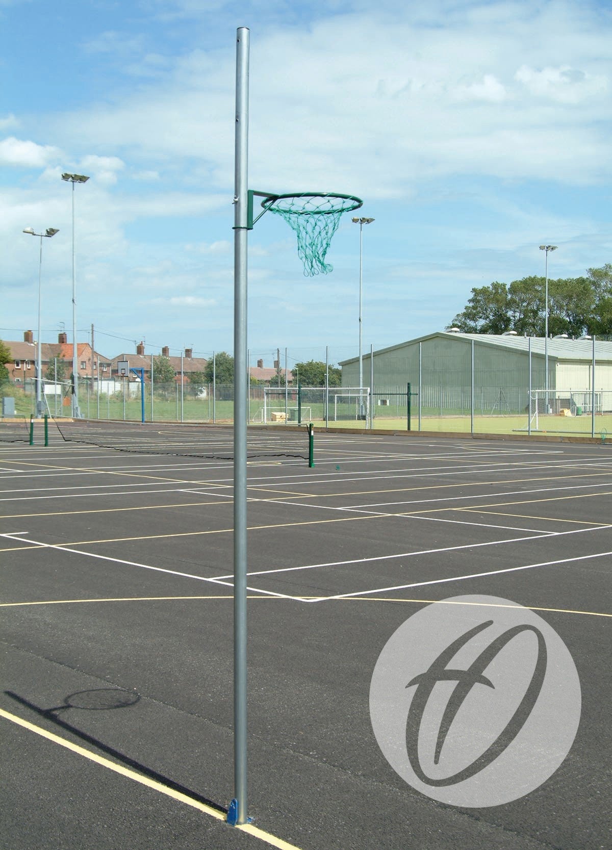 A Harrod Sport Regulation Netball Post with a socketed 16MM ring stands on an empty outdoor court, marked for play. Fences, buildings, and trees appear in the background beneath a partly cloudy sky.