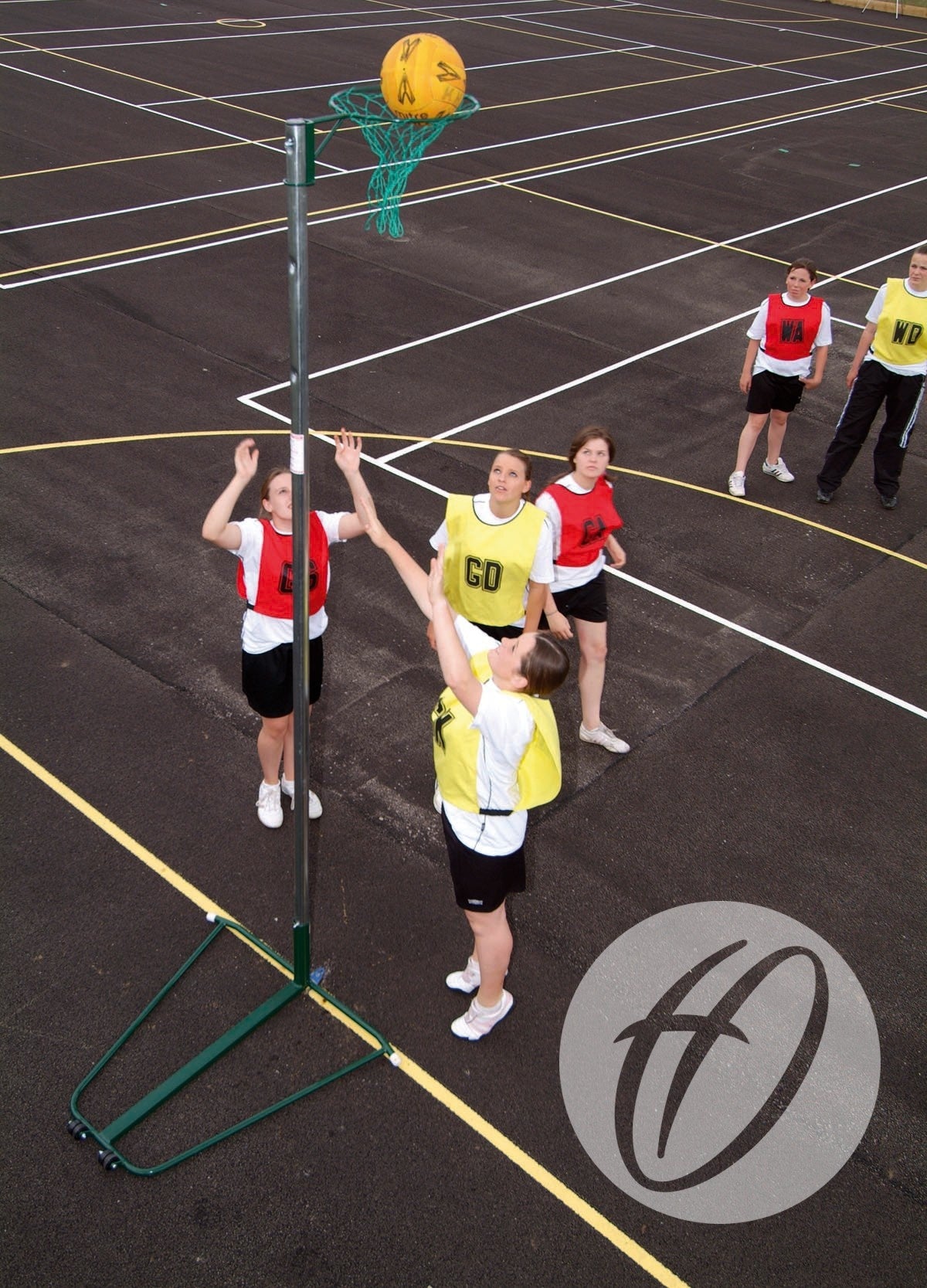 A group of women play netball on an outdoor court with the Harrod Sport NBL-023 Wheelaway Netball Post and Base (10mm Ring); a player in yellow bib shoots while others in yellow and red bibs watch—ideal for school use.