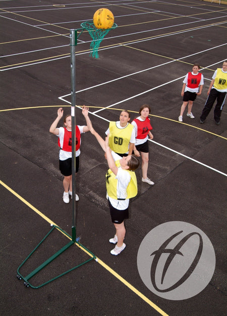 A group of women play netball on an outdoor court with the Harrod Sport NBL-023 Wheelaway Netball Post and Base (10mm Ring); a player in yellow bib shoots while others in yellow and red bibs watch—ideal for school use.