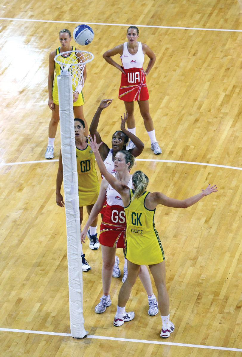 A netball match unfolds on a wooden court as a red team player aims to shoot the ball above Harrod Sport International Netball Posts - Socketed, while two yellow defenders attempt a block and others watch nearby.