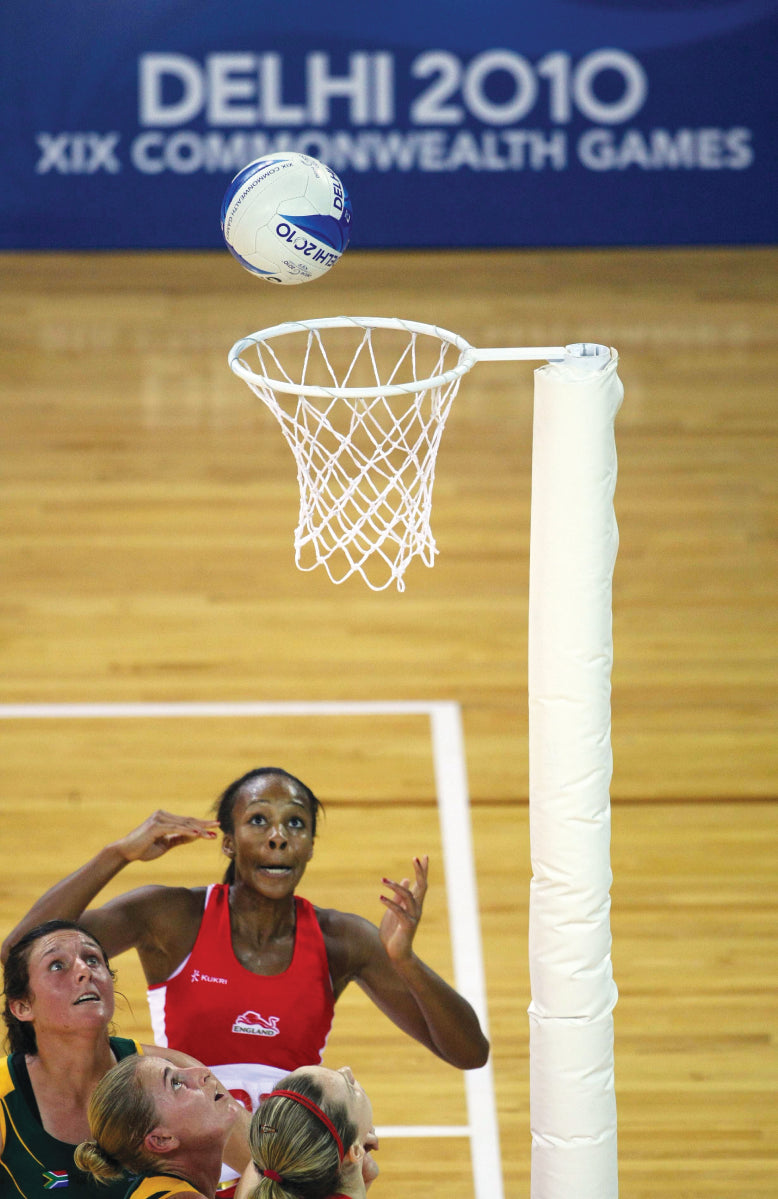 Four netball players watch a ball near the hoop with Harrod Sport International Netball Posts - Socketed during a 2010 Commonwealth Games match in Delhi, as a DELHI 2010 XIX COMMONWEALTH GAMES sign appears in the background.