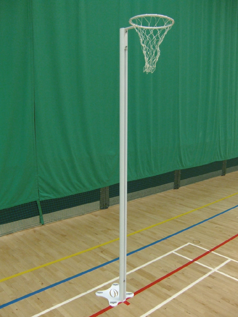 The Harrod Sport International Netball Posts - Floor Fixed feature reinforced aluminium uprights for stability. A white pole and net stand on a wooden indoor court, accented by colorful lines and a green curtain backdrop.