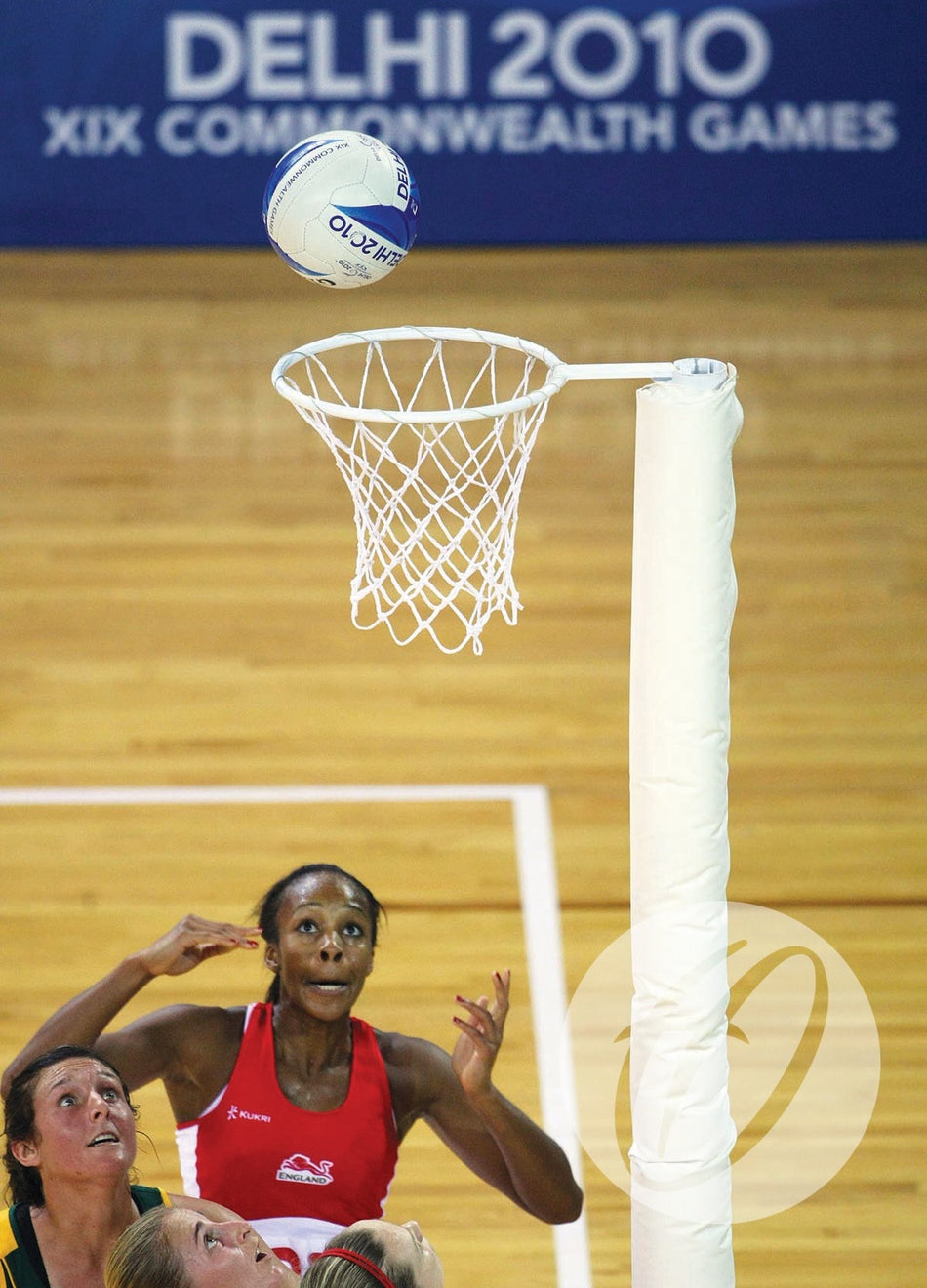 A netball player in a red uniform watches as the ball nears the Harrod Sport Competition White Netball Nets (Pair) during a Delhi 2010 Commonwealth Games match, with the wooden court and blue event banner in the background.