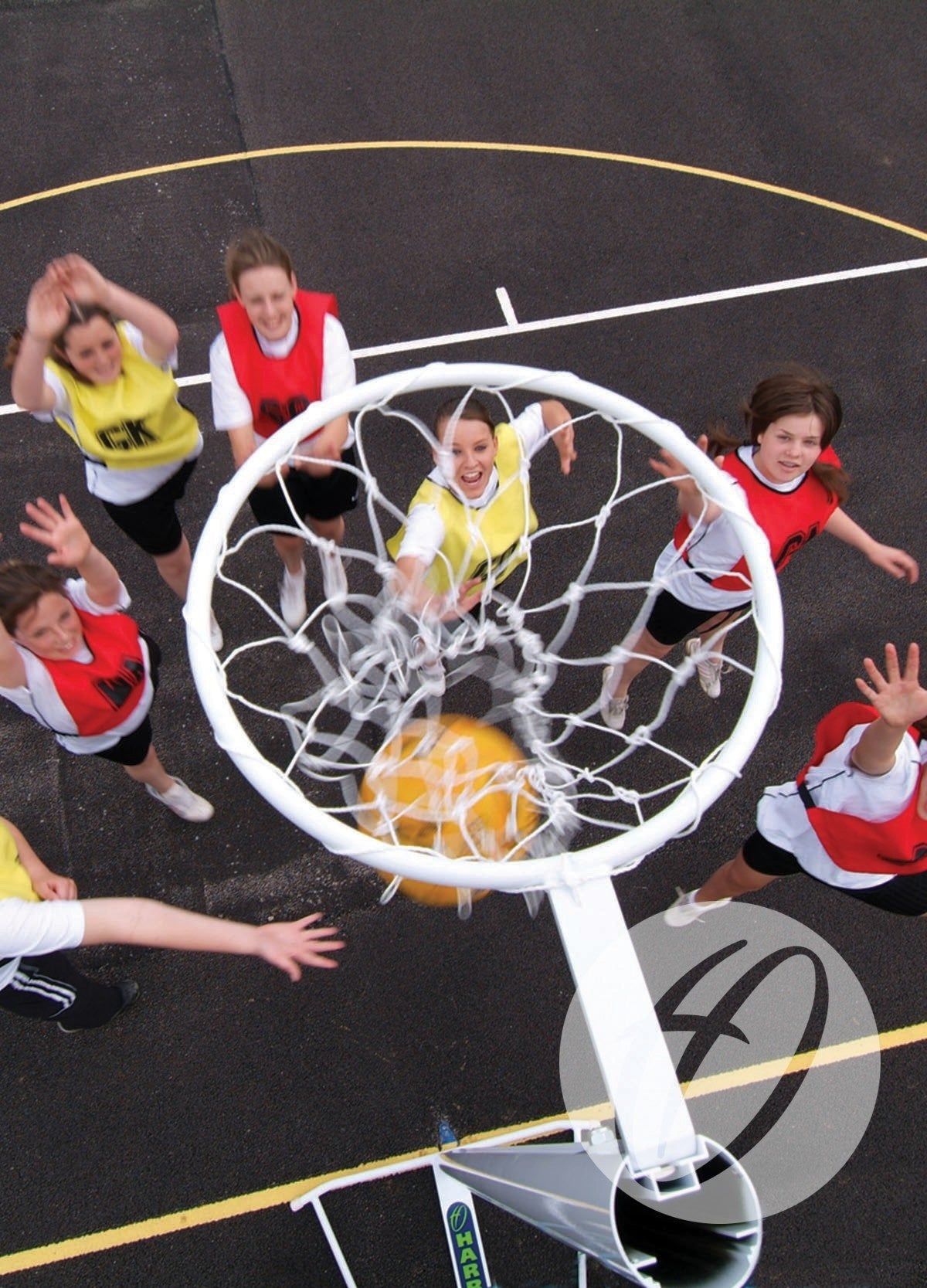An overhead view of young girls playing netball on an outdoor court with visible lines, reaching for the yellow ball as it goes through a Harrod Sport International Netball Post - Freestanding. Players wear red or yellow bibs.