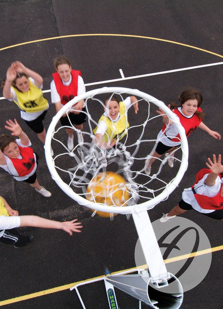An overhead view of young girls playing netball on an outdoor court with visible lines, reaching for the yellow ball as it goes through a Harrod Sport International Netball Post - Freestanding. Players wear red or yellow bibs.