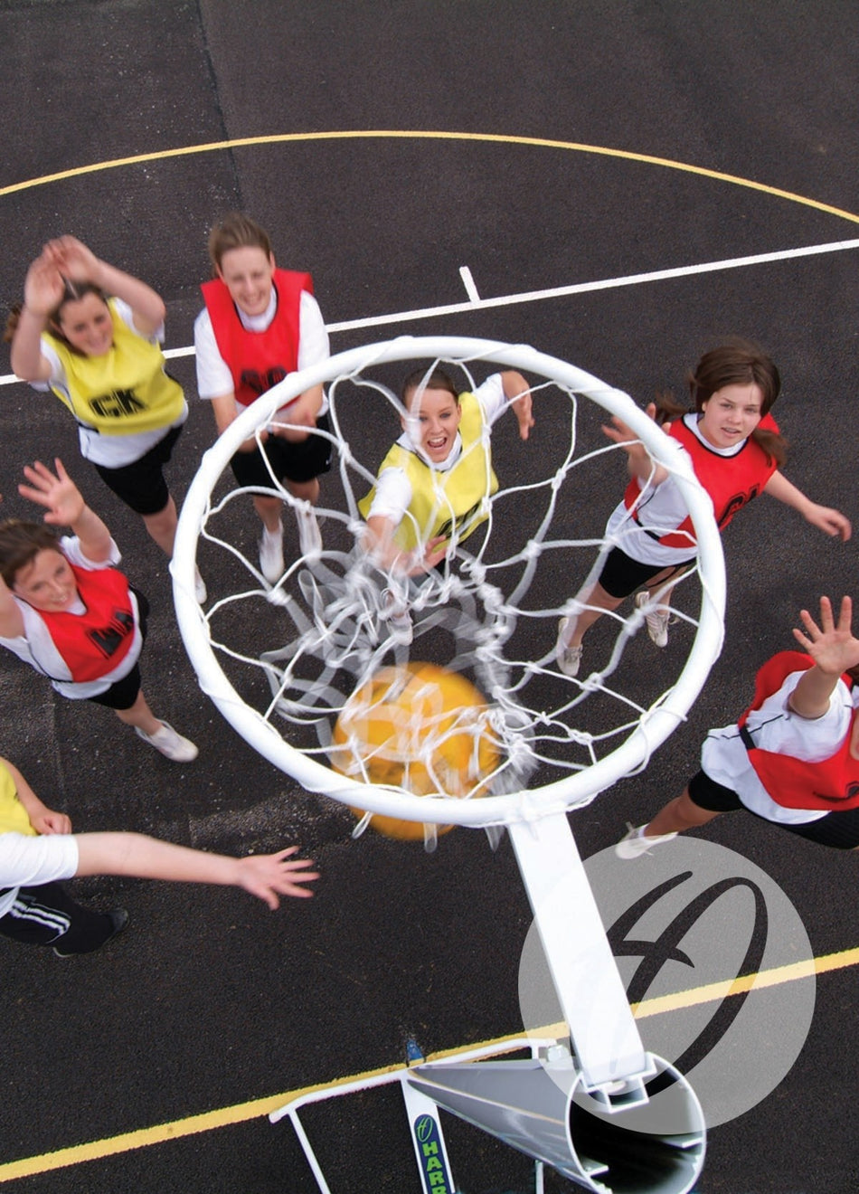 An overhead view of young girls playing netball on an outdoor court with visible lines, reaching for the yellow ball as it goes through a Harrod Sport International Netball Post - Freestanding. Players wear red or yellow bibs.