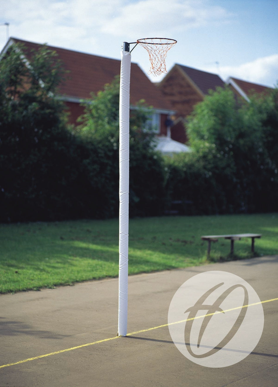 A Harrod Sport Standard Netball Protector in white covers the post on an outdoor court near a yellow line, with green grass, bushes, red-roofed houses in the background, and a wooden bench to the side.