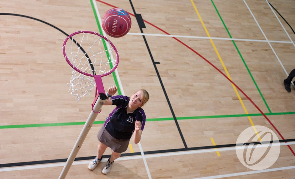 A woman in a purple sports uniform gets ready to catch a red netball near Harrod Sport Pink Netball Posts—freestanding with a 16MM ring—on an indoor court with colorful boundary lines.