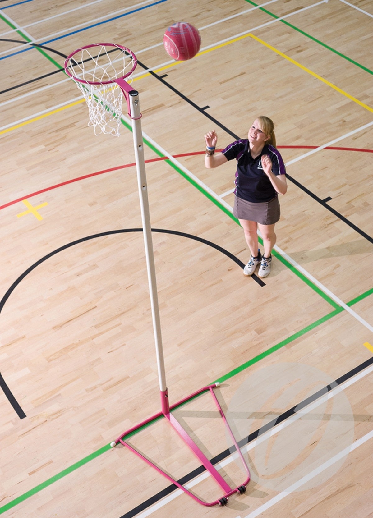 A woman plays netball indoors, jumping to shoot a pink ball at the Harrod Sport Single Pink Netball Post—Freestanding with a 10MM ring—on a wooden court marked with colorful lines.