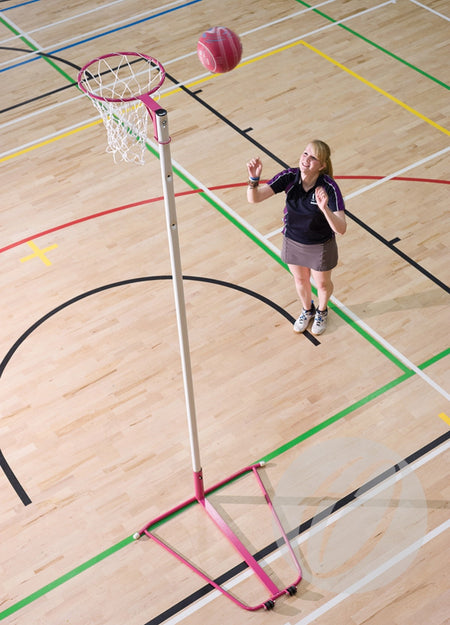 A woman plays netball indoors, jumping to shoot a pink ball at the Harrod Sport Single Pink Netball Post—Freestanding with a 10MM ring—on a wooden court marked with colorful lines.