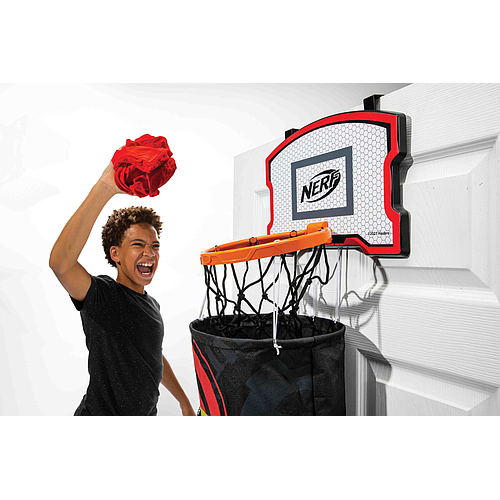 A young person excitedly tosses a rolled-up red shirt into the Nerf 3-In-1 Laundry Layup, an over-the-door hoop by Nerf mounted on a white door, celebrating after making the shot.