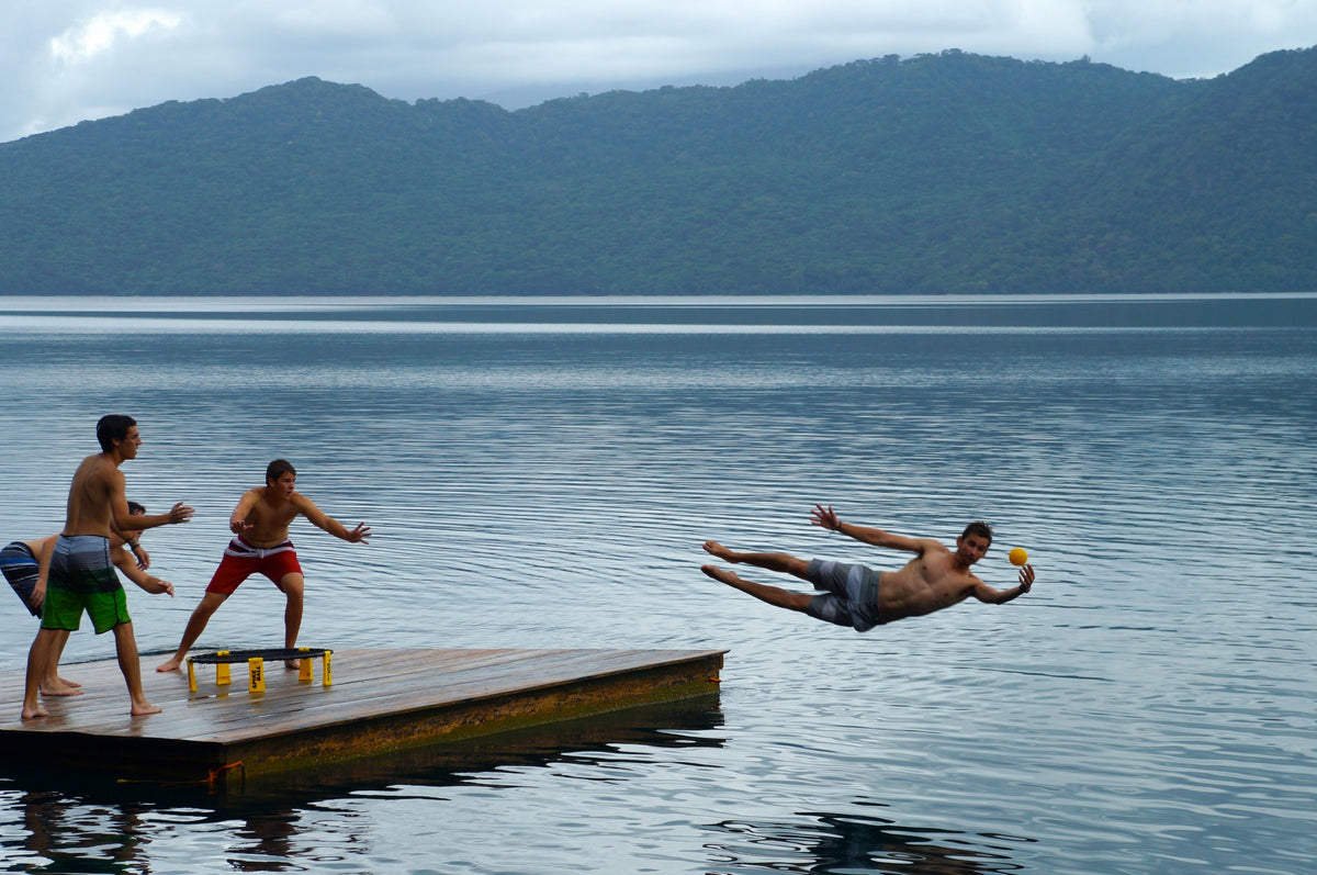 Three people play Spikeball with the Kankakee Spikeball Standard Set on a wooden dock by a lake; one dives mid-air with a yellow cup as others reach out, surrounded by forested hills and cloudy skies in this energetic outdoor scene.