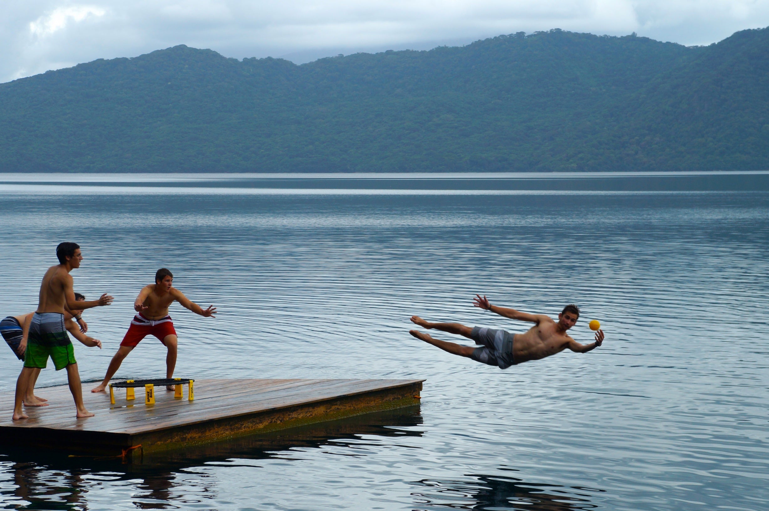 Three people play Spikeball with the Kankakee Spikeball Standard Set on a wooden dock by a lake; one dives mid-air with a yellow cup as others reach out, surrounded by forested hills and cloudy skies in this energetic outdoor scene.