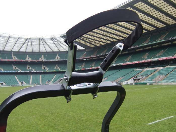 Close-up of a scrum machine’s padded arm and handle on a rugby field, with rugby posts, empty stadium seating, and the Harrod Sport Hinged Post Assembly Roller in the background.