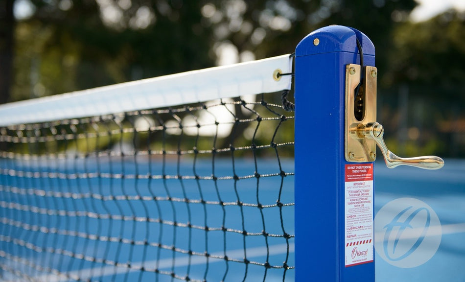 Close-up of the Harrod Sport Mini Tennis Net - P17 post with a blue and gold tension adjuster on an outdoor court, showing the net and blue surface in the background.
