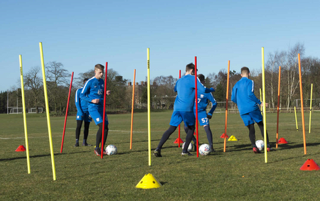 Four soccer players in blue uniforms practice dribbling and agility on a grassy field, weaving around Samba Sports' 1.7m Boundary Poles (set of 12) that mark boundaries, with soccer balls at their feet beneath a clear blue sky.