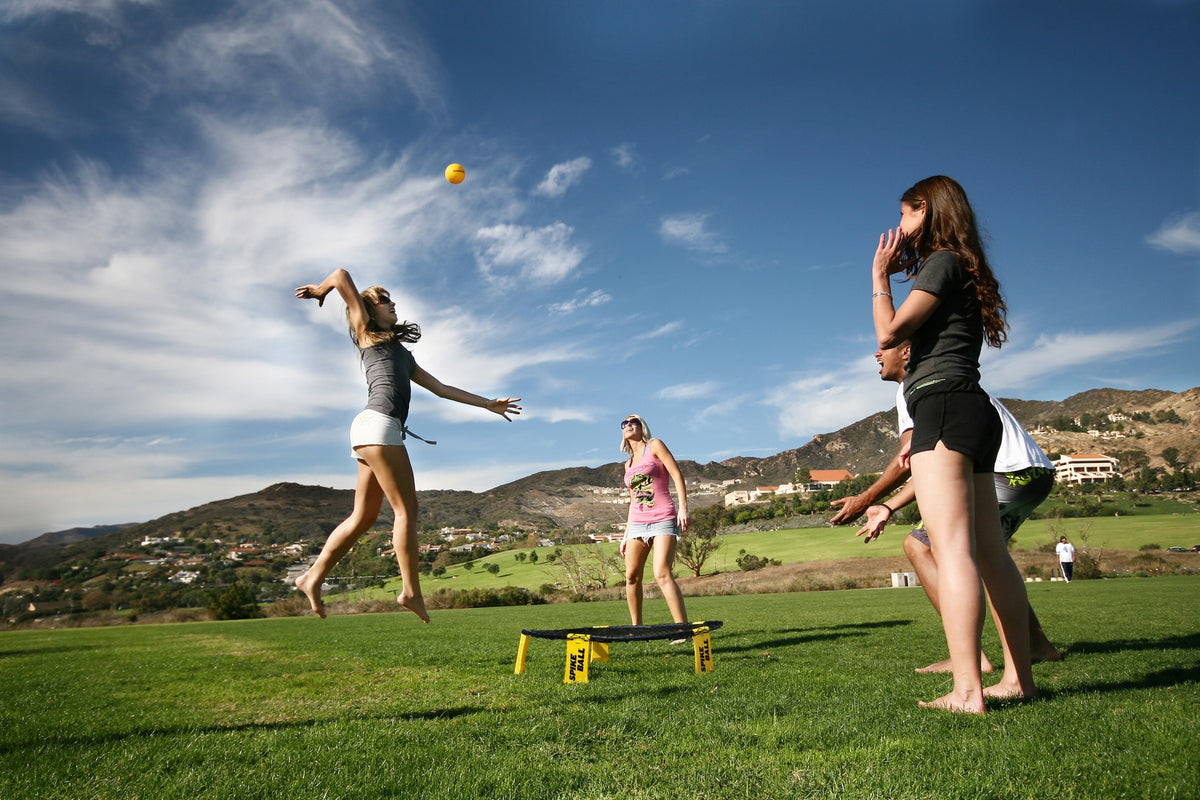 Four young people play Kankakee Spikeball's Spikeball Standard Set on a grassy field under a blue sky. One player jumps to hit the ball over the adjustable net while others get ready. Hills and houses can be seen in the background.