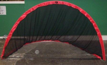 The Hunts County Pop up Backstop/Goal features a red frame and black netting, standing on a concrete floor near a green wall—perfect for training sessions and easy to transport in its included carry bag.