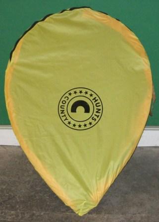 A yellow, leaf-shaped Hunts County Pop up Backstop/Goal with a black circular Hunts County logo in the center, standing upright against a green wall and grey floor.
