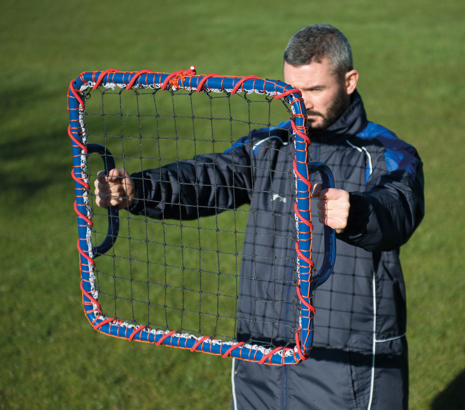 A person on grass holds the Precision Hand-Held Rebounder by Precision Training, featuring a blue and red frame with black mesh and handles—ideal for sports practice and reaction training. The person wears a dark jacket.